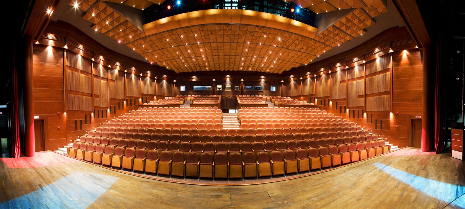 An empty concert hall with rows of orange seats facing a wooden stage under warm lights