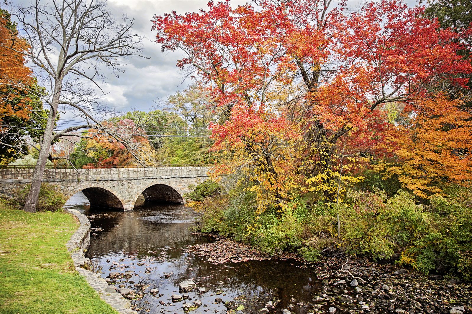 A stone arch footbridge over a shallow creek under red and orange autumn trees in a city park